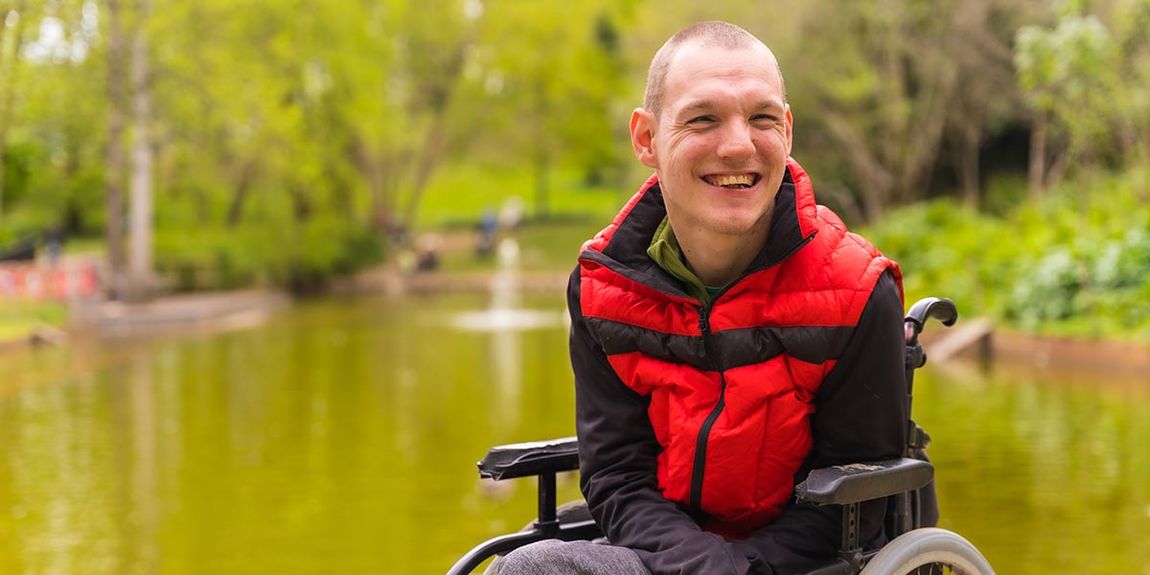 Ein junger Mann mt roter Jacke im Rollstuhl sitzend lächelt fröhlich in die Kamera.