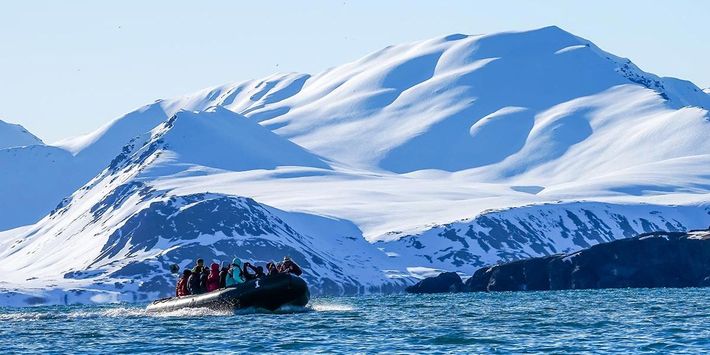 Eine Gruppe Menschen fährt in einem Zodiac Boot vor einem schneebedeckten Berg im Magdalenenfjord.