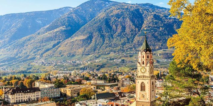 Herbstlicher Blick über die Altstadt von Meran mit Stadtturm, bunten Bäumen und der umliegenden Berglandschaft Südtirols im goldenen Herbstlicht.