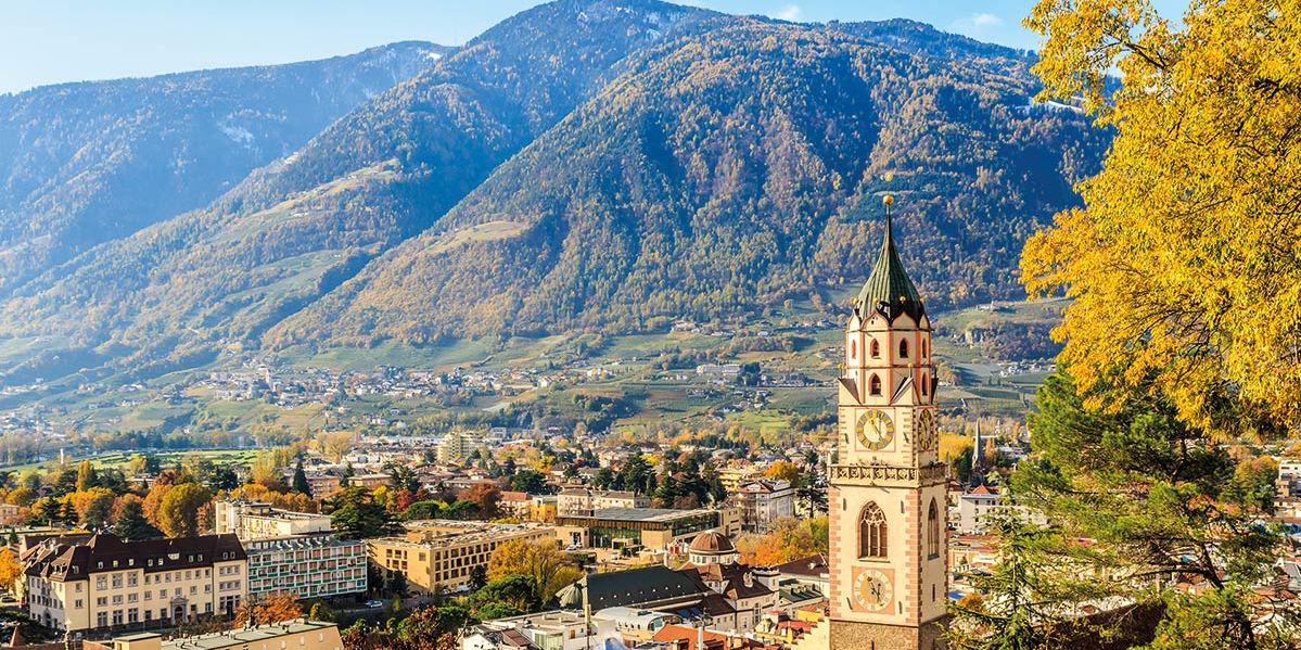 Herbstlicher Blick über die Altstadt von Meran mit Stadtturm, bunten Bäumen und der umliegenden Berglandschaft Südtirols im goldenen Herbstlicht.