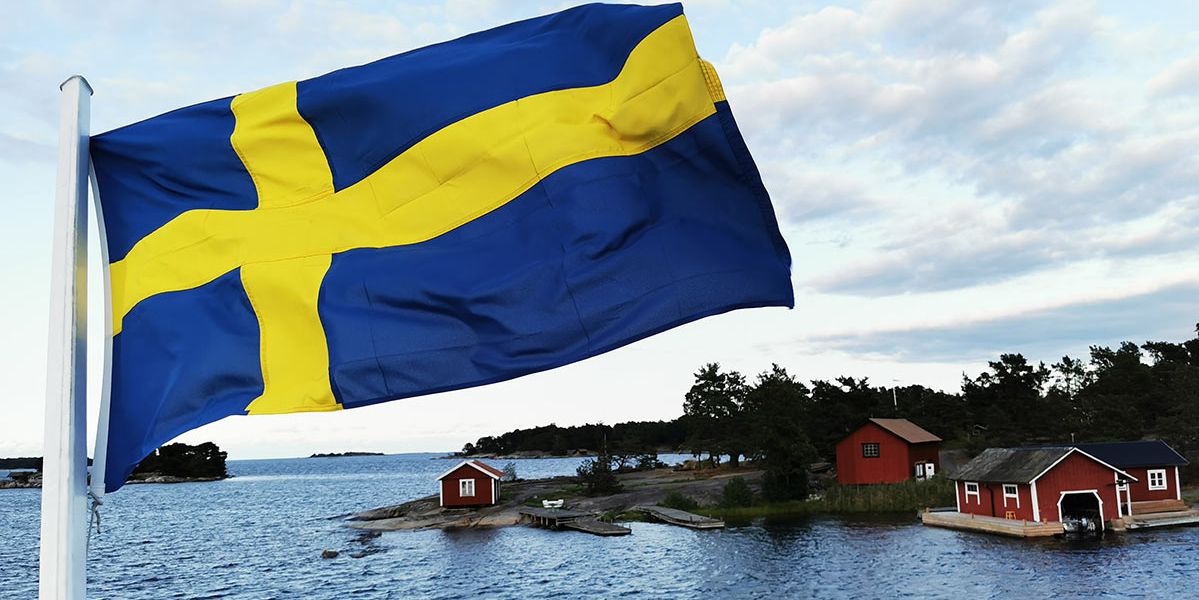 Die schwedische Flagge weht vor einem Fjord, mit einer Insel mit Blockhütten in Ochsenblutrot.