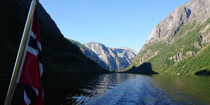 Die norwegische Flagge weht am Heck eines Schiffes mit Blick in einen Fjord mit steilen Felswänden und blauem Wasser.