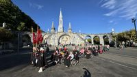 Pilgerprozession mit Rollstuhlfahrern vor der Rosenkranzbasilika in Lourdes, Frankreich, auf dem Vorplatz des Wallfahrtsheiligtums bei blauem Himmel.