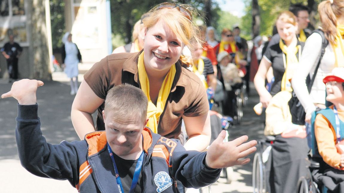 Eine ehrenamtliche Helferin schiebt einen im Rollstuhl sitzenden Jungen fröhlich durch Lourdes.