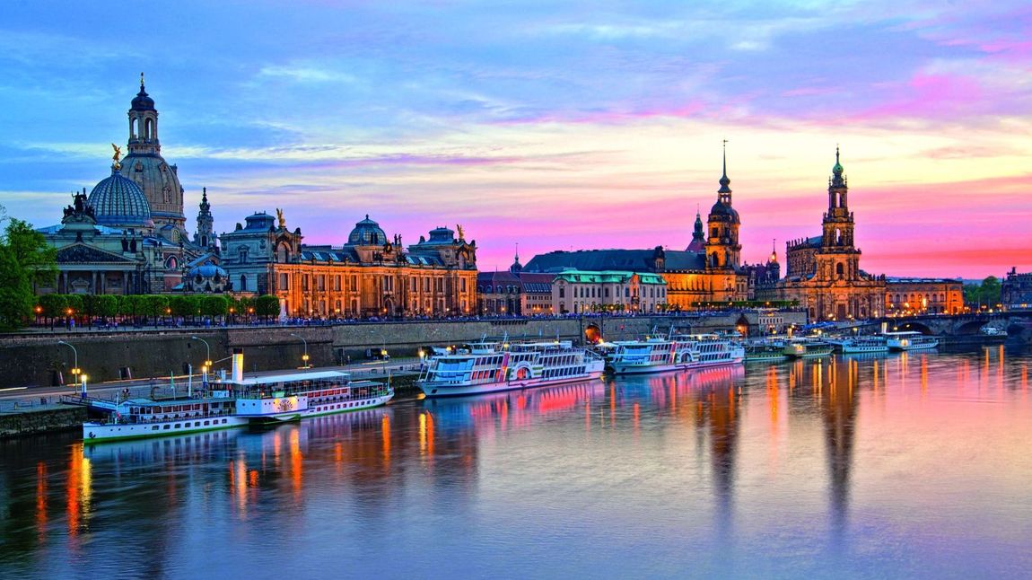 Viele Flusskreuzfahrtschiffe liegen auf der Elbe vor Dresden, die Skyline mit Frauenkirche und Hofkirche leuchtet im Sonnenuntergang.