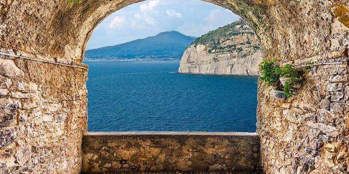 Blick durch eine gemauerte Fensternische auf das balaue Meer des Golfs von Neapel mit dem Vesuv im Hintergrund.