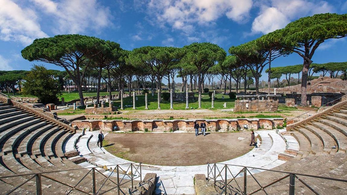 Blick von oben in das Amphitheater in Ostia Antica mit vielen Tribünenplätzen vor Pinienbäumen und blauem Himmel.