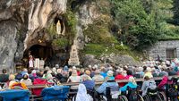 Eucharistiefeier an der Grotte von Massabielle in Lourdes mit zahlreichen Pilgerinnen und Pilgern, darunter Menschen im Rollstuhl, während ein Priester am Altar vor der Marienstatue zelebriert.
