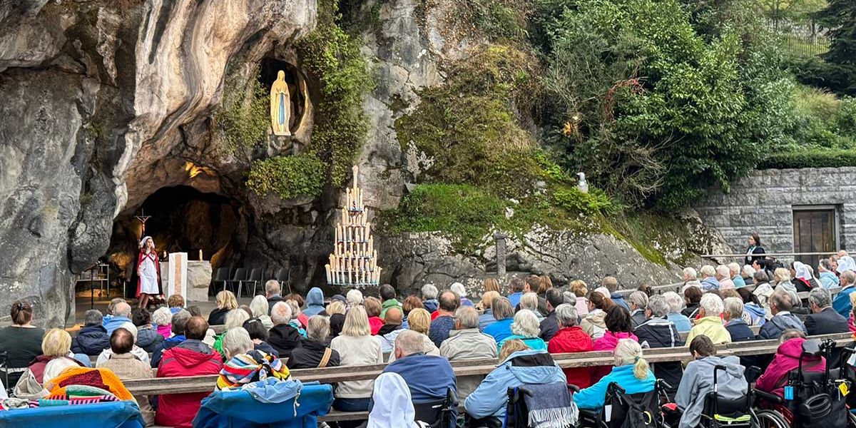 Eucharistiefeier an der Grotte von Massabielle in Lourdes mit zahlreichen Pilgerinnen und Pilgern, darunter Menschen im Rollstuhl, während ein Priester am Altar vor der Marienstatue zelebriert.