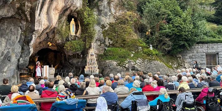 Eucharistiefeier an der Grotte von Massabielle in Lourdes mit zahlreichen Pilgerinnen und Pilgern, darunter Menschen im Rollstuhl, während ein Priester am Altar vor der Marienstatue zelebriert.